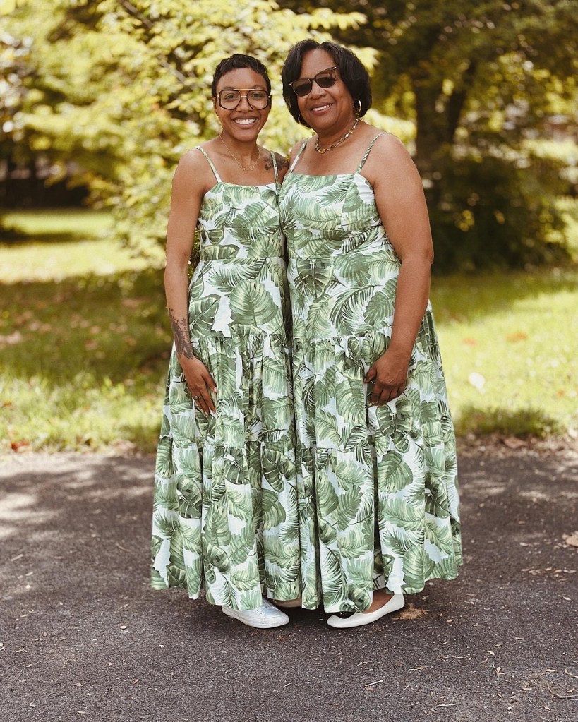 Mother and daughter smiling and posing together outdoors, both wearing matching green dresses with leaf patterns.