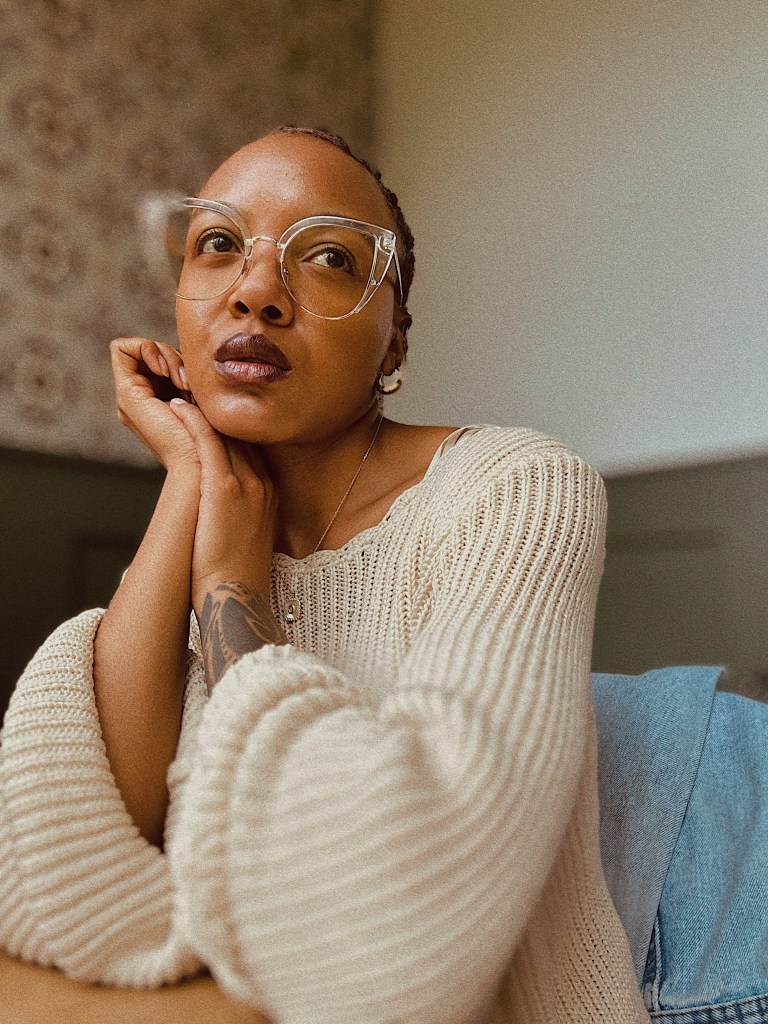 Courtney Elayne, a woman with short hair wearing clear glasses and a cream-colored knit sweater, sitting with their chin resting on their hand, against a softly patterned background.