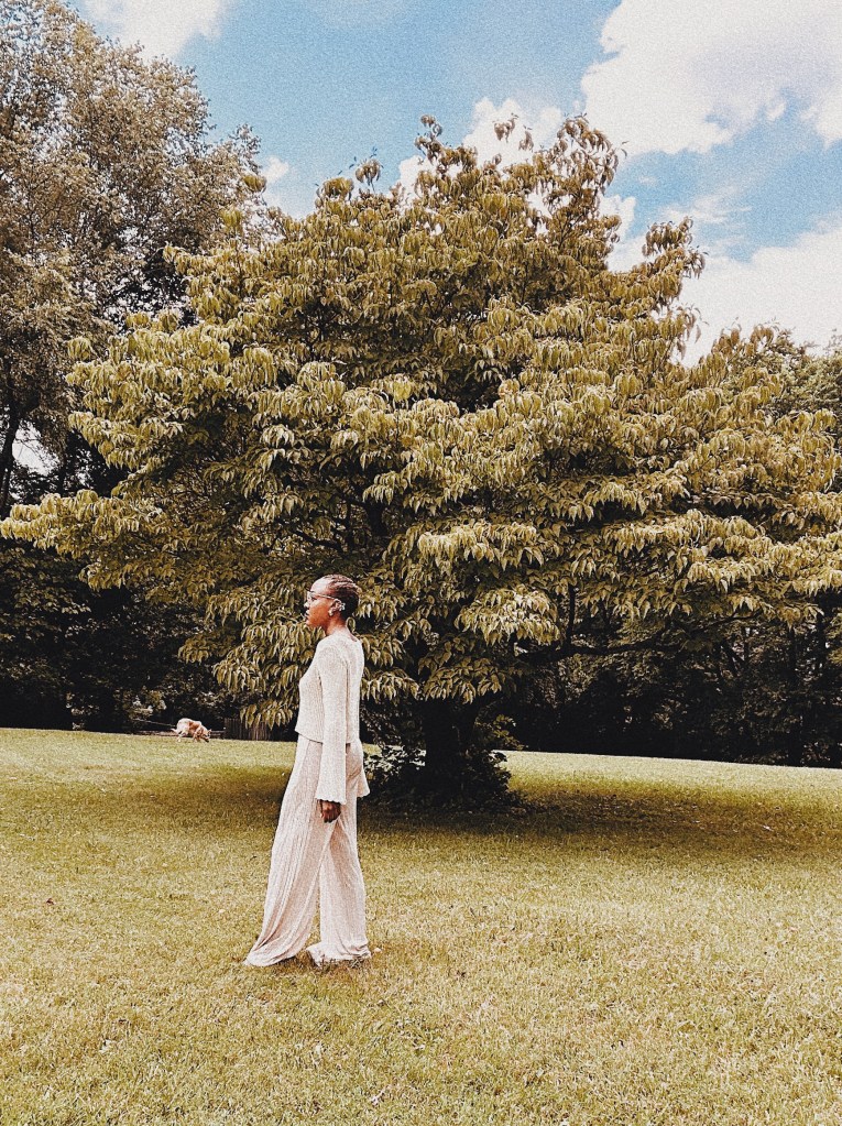 Courtney Elayne, a woman in a flowing cream outfit stands in her backyard, grounded in nature and reflection. Behind her, a large leafy tree symbolizes growth, while the open sky above suggests clarity and openness. This image reflects the quiet, thoughtful beginning of meaningful writing and understanding the reader.