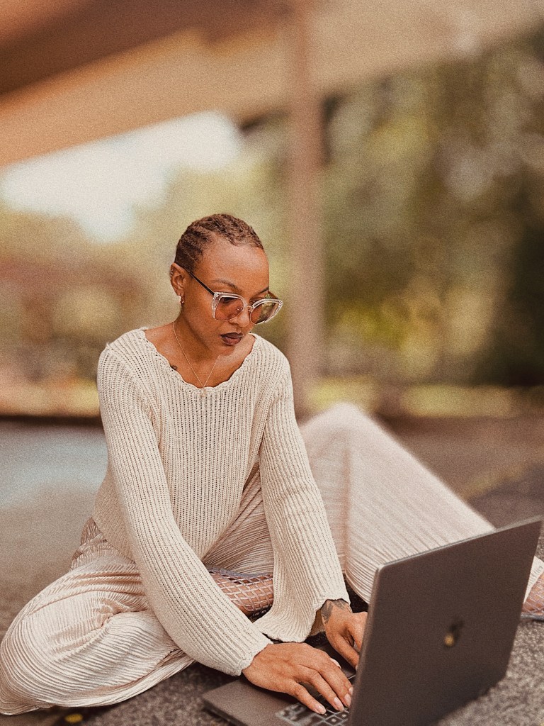 Courtney Elayne, a woman sitting outdoors, wearing a beige sweater and large glasses, working on a laptop. The background features blurred greenery and soft lighting.